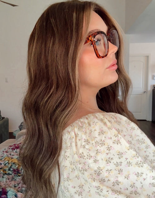 A woman with long, wavy brown hair and brown cat-eye eyeglasses, wearing a cream floral off-the-shoulder top, shown in profile in an indoor setting.