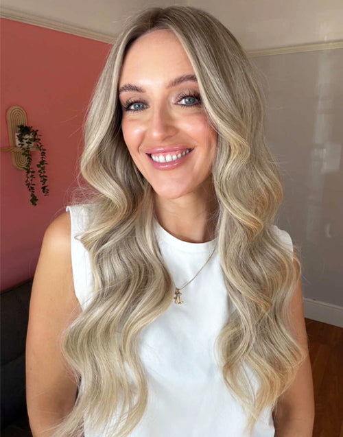 Real-life home photo of a smiling woman wearing a long wavy ash blonde balayage human hair wig