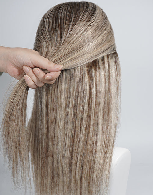 A close-up of a long, straight wig or hair extension piece shown from the back, with a hand lifting a section of the hair to demonstrate texture and layering. The color is a blend of light brown and blonde highlights.