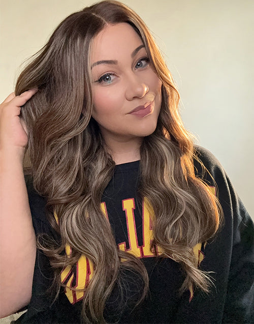 Woman running fingers through long wavy wig with honey blonde balayage highlights showing natural scalp parting and soft hair texture.