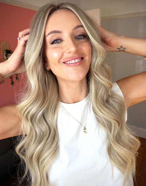 Close-up front view of a smiling woman styling a long wavy ash blonde balayage human hair wig with her hands