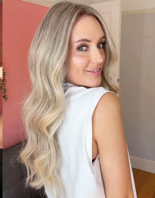Side-back view of a smiling woman wearing a long wavy ash blonde balayage human hair wig